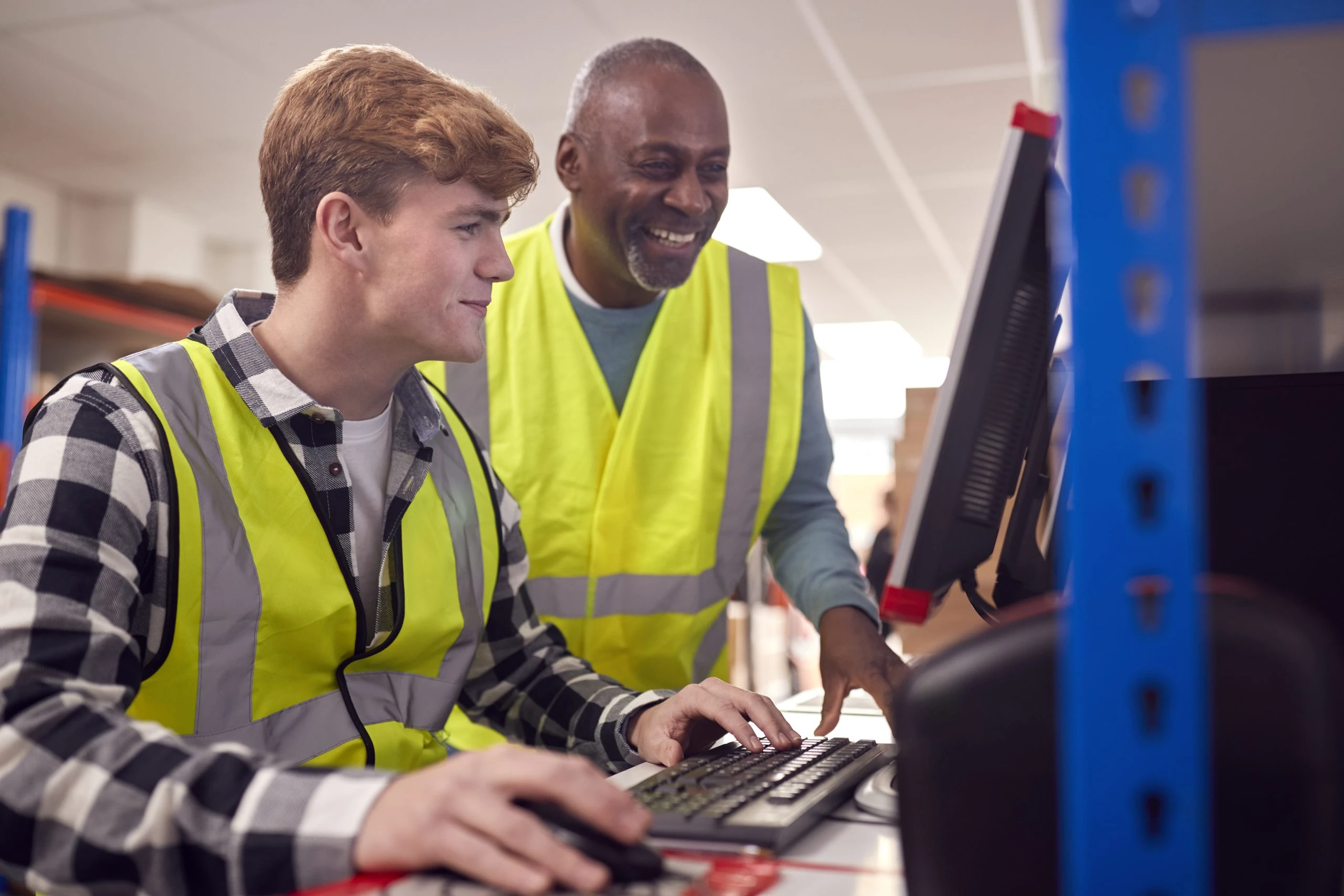 Male Intern With Supervisor Working In Busy Modern Warehouse On Computer Terminal